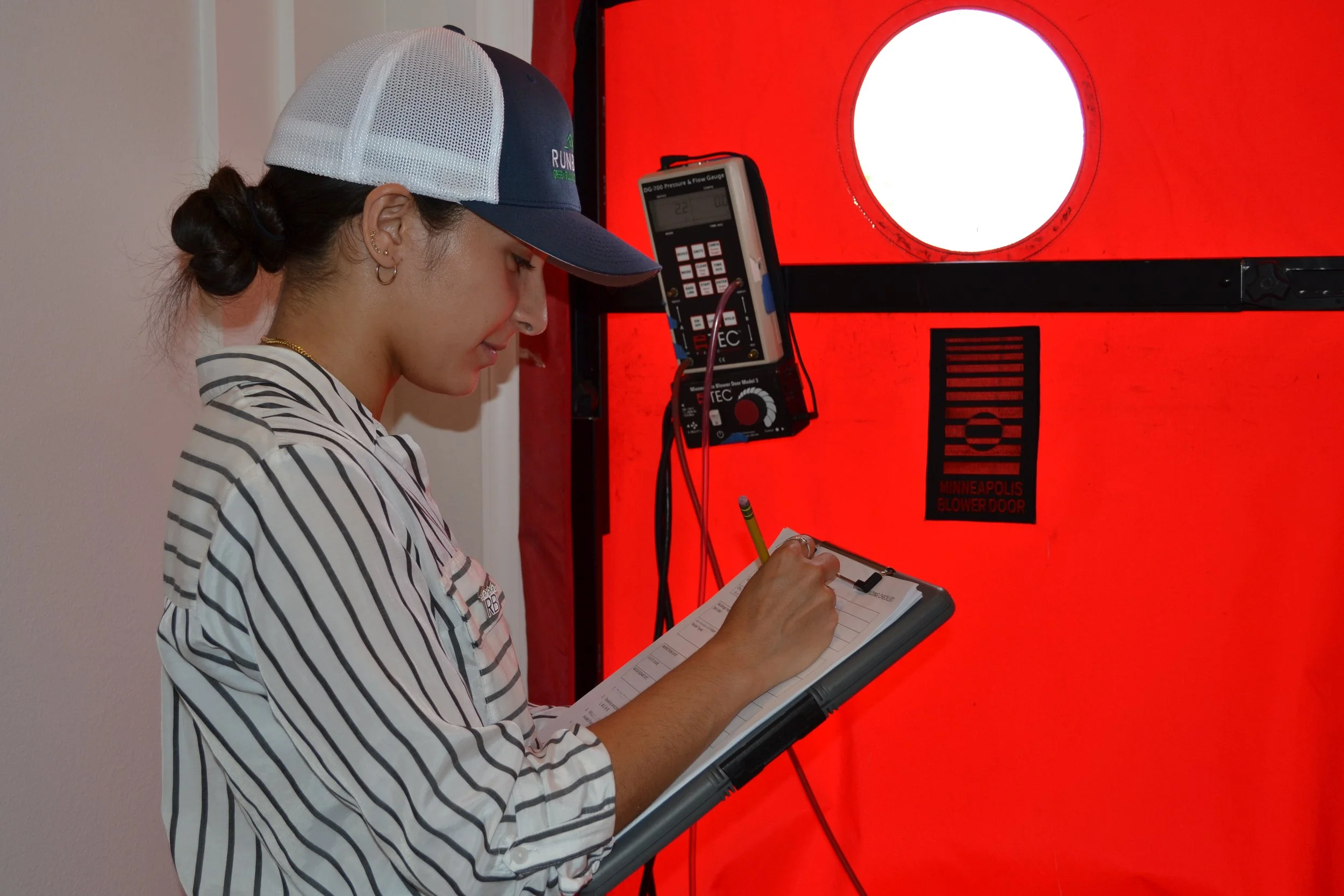 A woman wearing a gray and white trucker hat and striped shirt is writing in a notepad while standing in front of a red blower door test setup with a digital pressure and flow gauge.