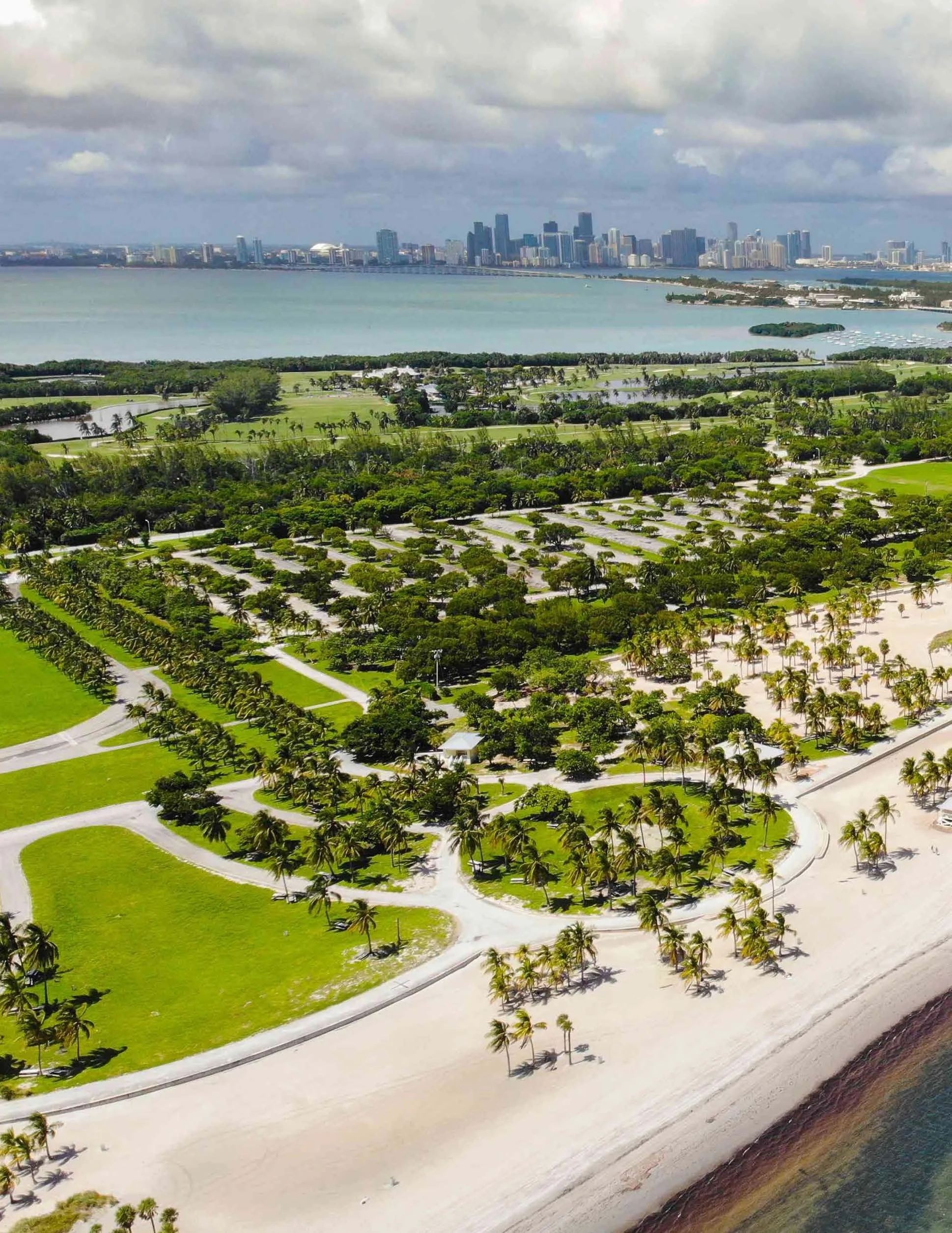 Aerial view of a golf course with palm trees, white sand bunkers, and lush green fairways, with a large body of water; in the background, the Miami skyline with high-rise buildings under a cloudy sky.