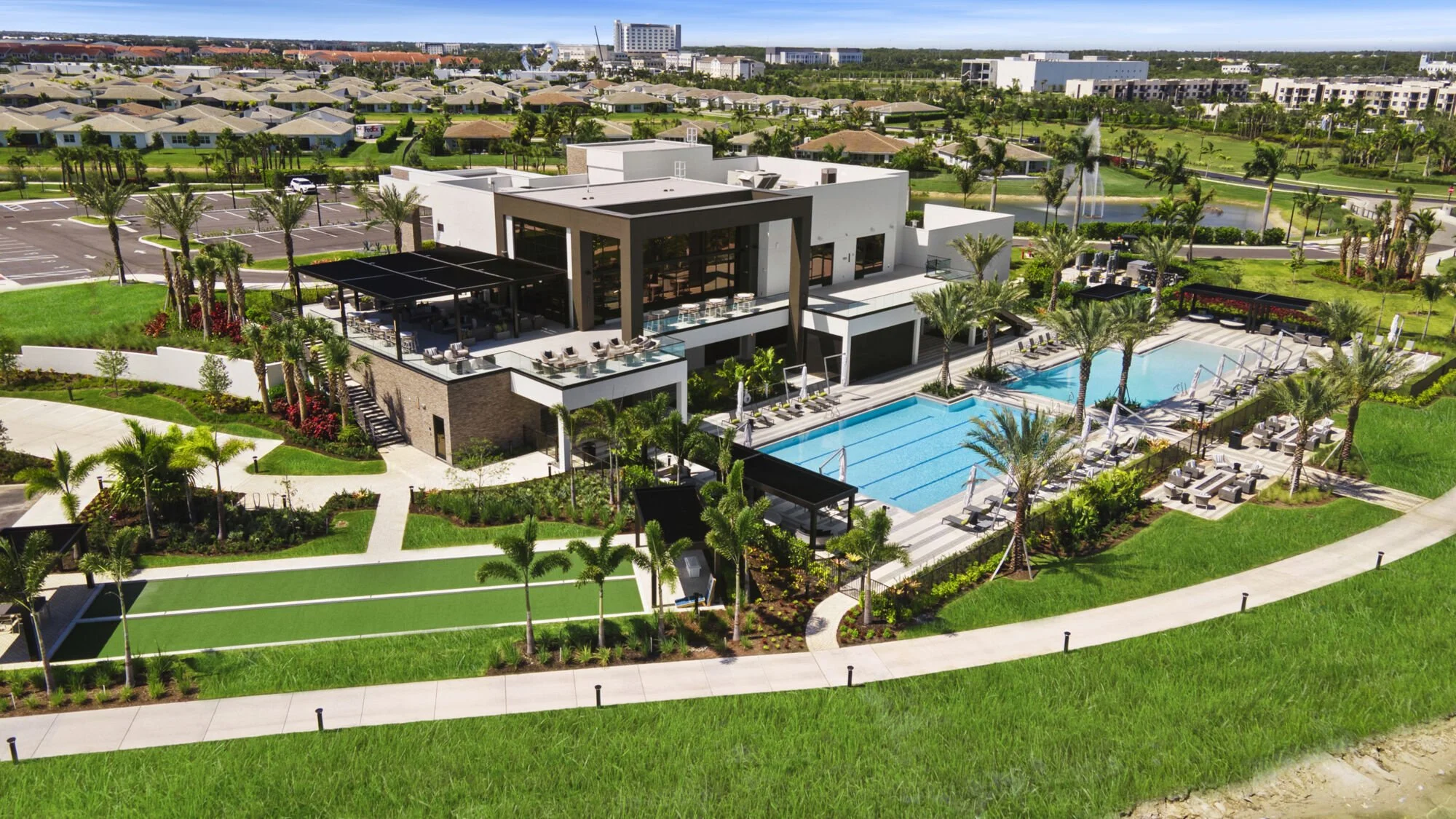 Aerial view of a modern residential complex with outdoor swimming pools, surrounded by palm trees, well-maintained lawns, and a parking lot, with city buildings in the background.