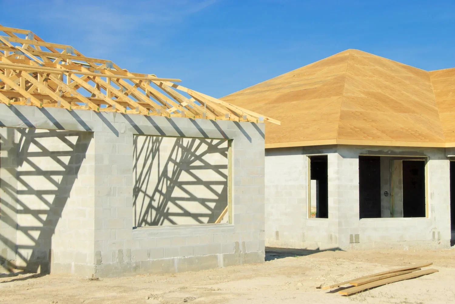 Construction site with partially built concrete houses, wooden roof trusses, and clear blue sky.