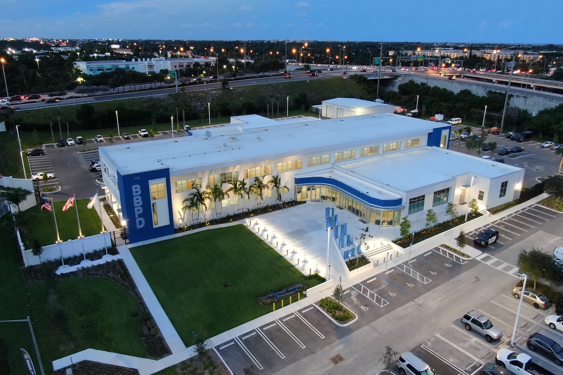 Aerial view of a modern building with blue and white exterior, palm trees, and a parking lot with some cars. In the background, a highway with moving cars and city lights at dusk.
