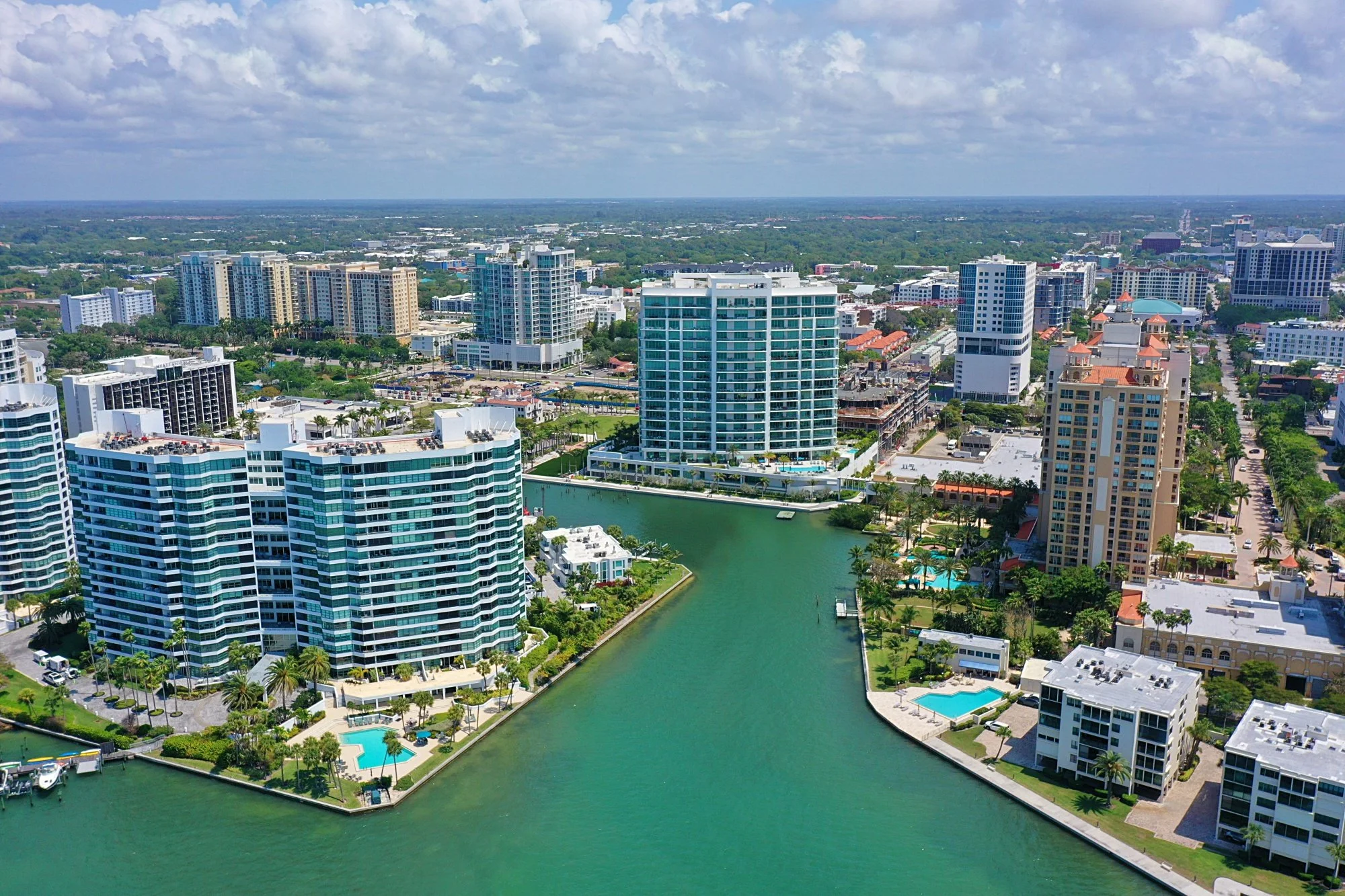 Aerial view of a city with modern high-rise buildings, a waterway, and lush greenery under a partly cloudy sky.