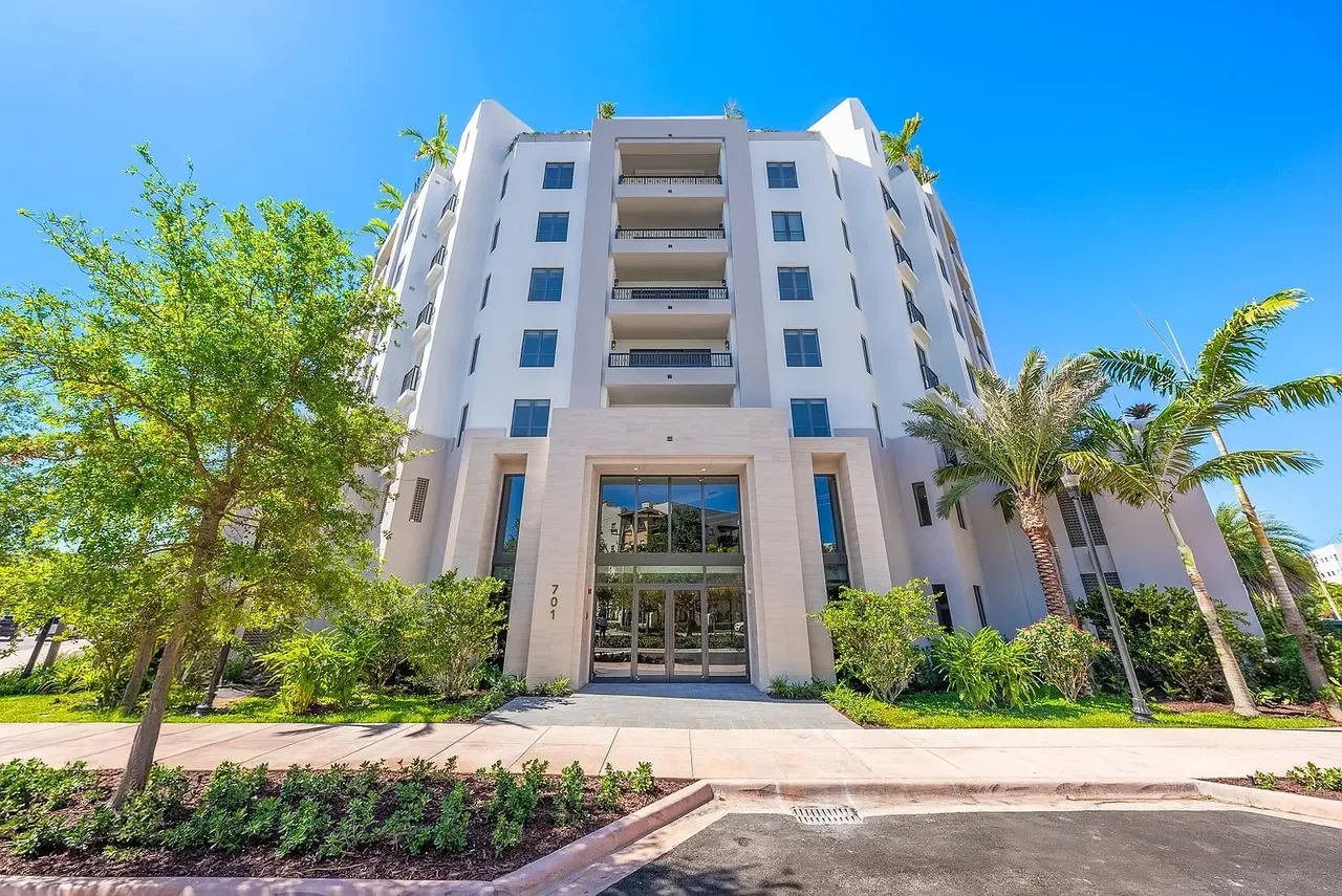 Modern white residential building with balconies, surrounded by palm trees and greenery, under a bright blue sky.