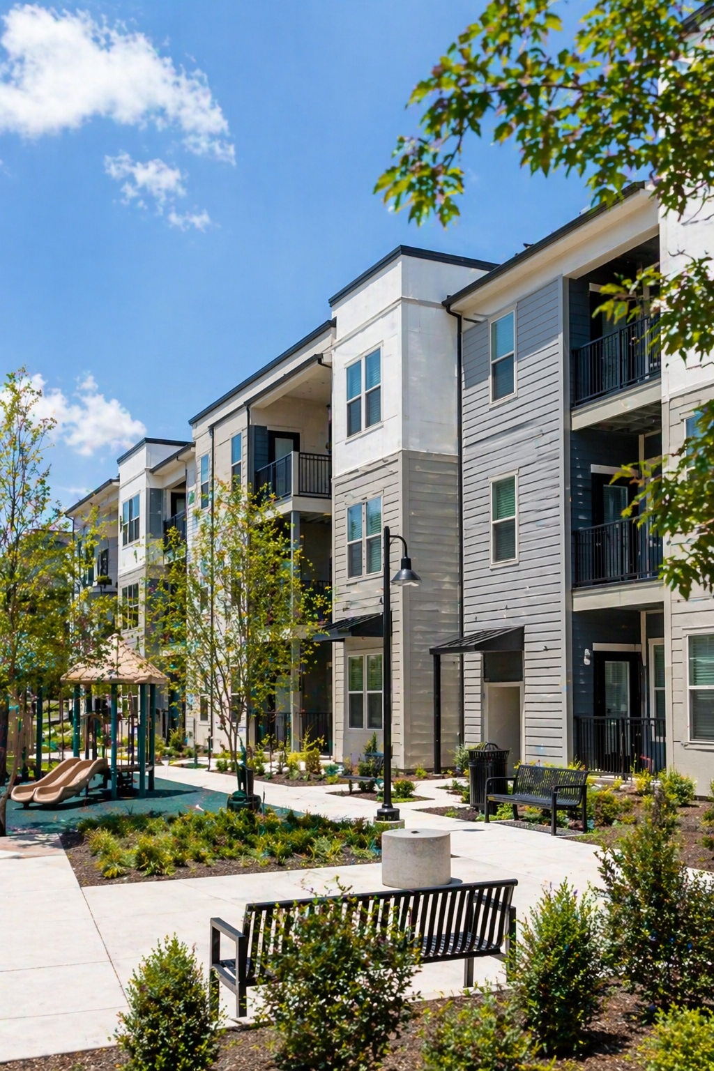 Modern apartment building with balconies, surrounded by a landscaped courtyard with trees, benches, and playground equipment under a blue sky.