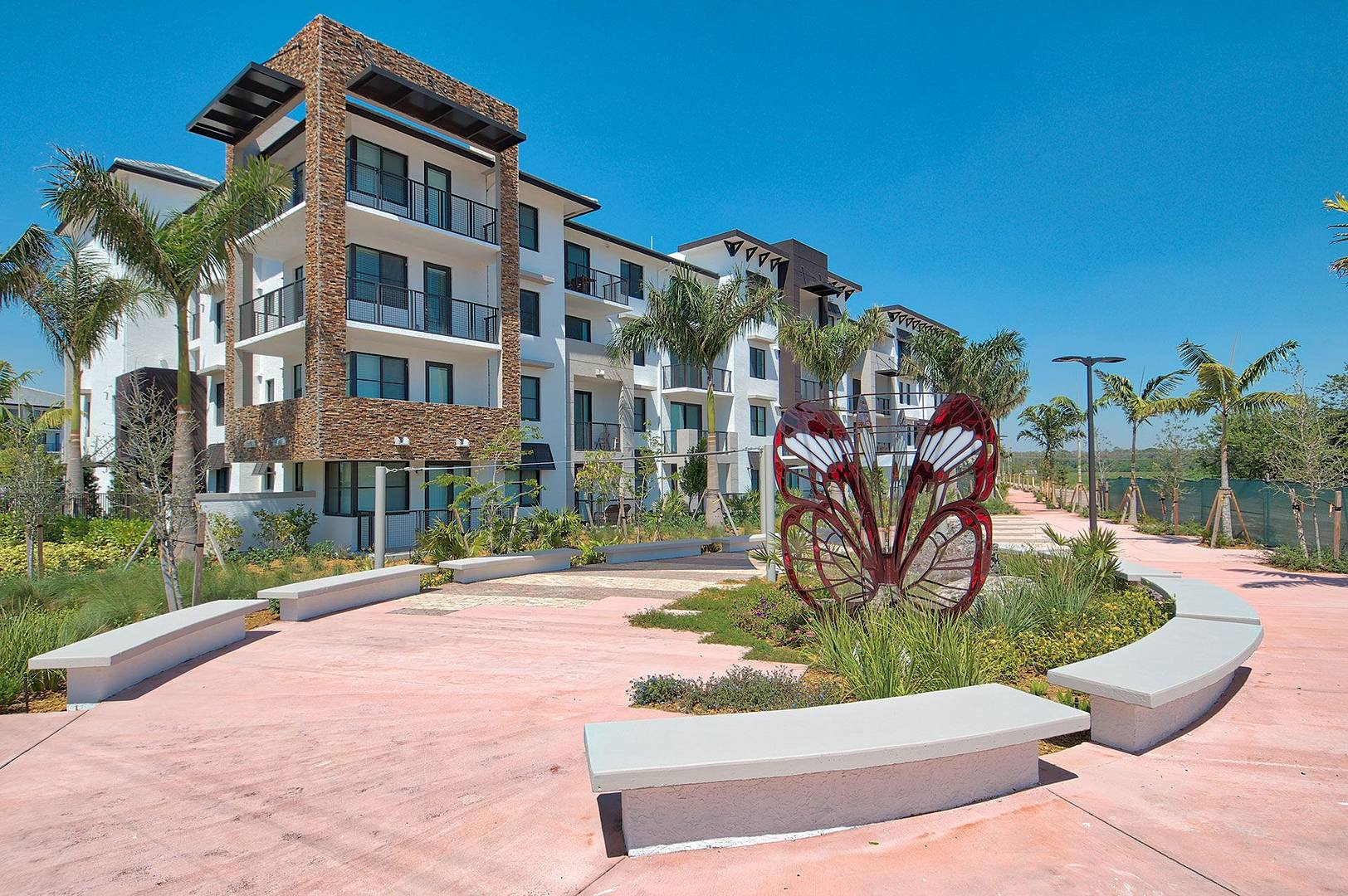 Modern apartment building with balconies, surrounded by palm trees and greenery, with a pink walking path and butterfly sculpture in the foreground under a clear blue sky.