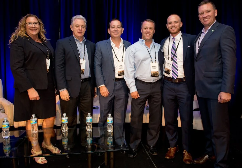 Six professionally dressed individuals standing together on a stage with a dark blue curtain background, smiling for the photo at a conference or event wearing name badges.