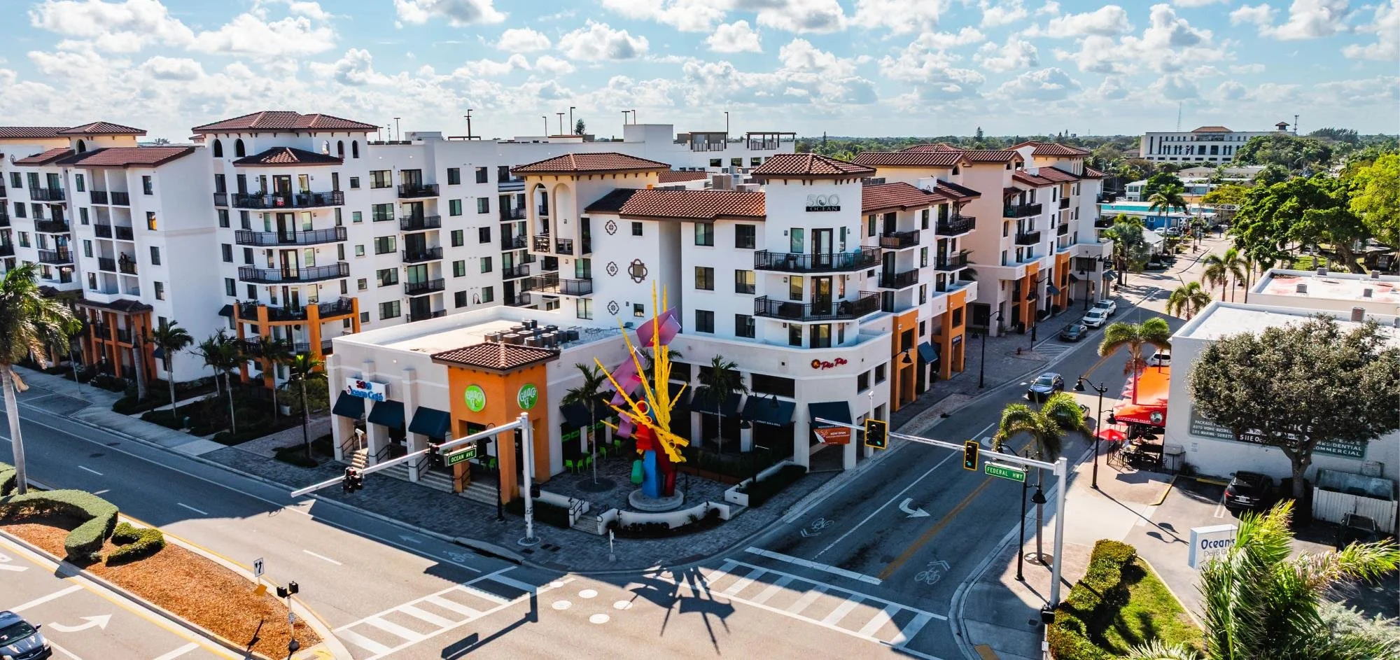 Aerial view of a multi-story shopping and residential complex with shops on the ground floor, surrounded by streets, palm trees, and parking lots, with a colorful sculpture at the corner of the building.