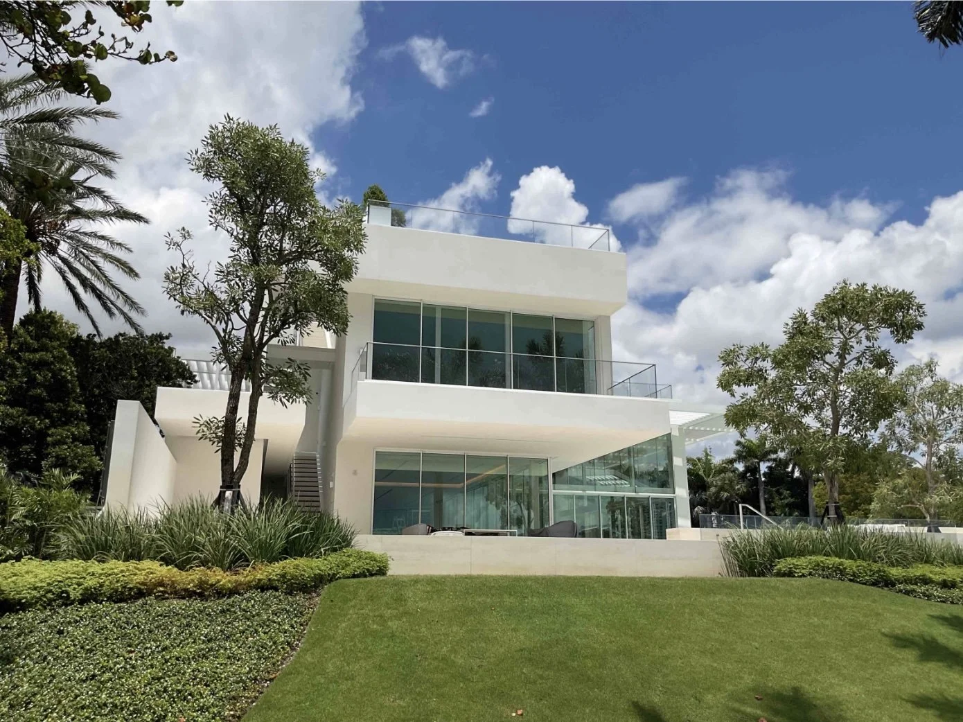 Modern white multi-story house with large glass windows and balconies, surrounded by lush green trees and grass, under a partly cloudy blue sky.