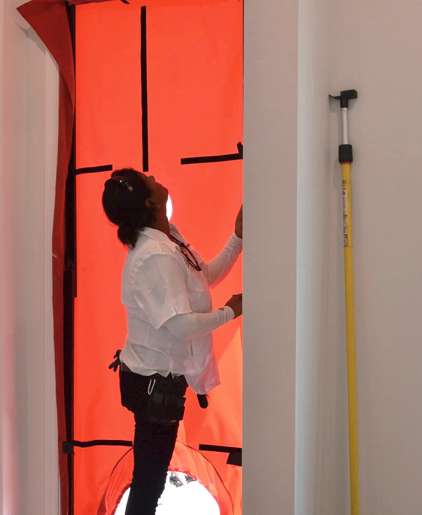 RunBrook performing a blower door test, includes an employee wearing a white shirt and black pants standing inside a bright red tunnel or inflatable structure, with a white wall on the right and a yellow-handled tool leaning against the wall.