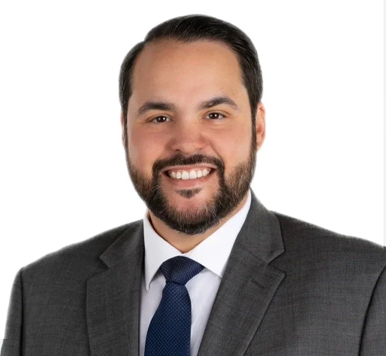 A man with dark hair, styled back, and a beard, wearing a gray suit, white shirt, and navy tie, smiling against a white background.
