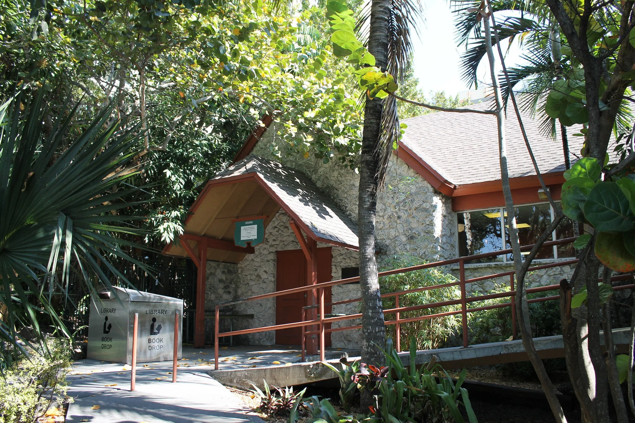 A stone building with a red-tiled roof surrounded by lush green trees and plants, with a sign indicating a book drop near the entrance, and a metal railing along a small pathway.