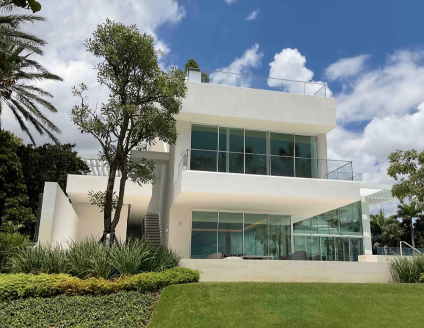 Modern white multi-story house with large windows, glass railings, and modern outdoor furniture, surrounded by landscaped greenery and trees, under a partly cloudy sky.