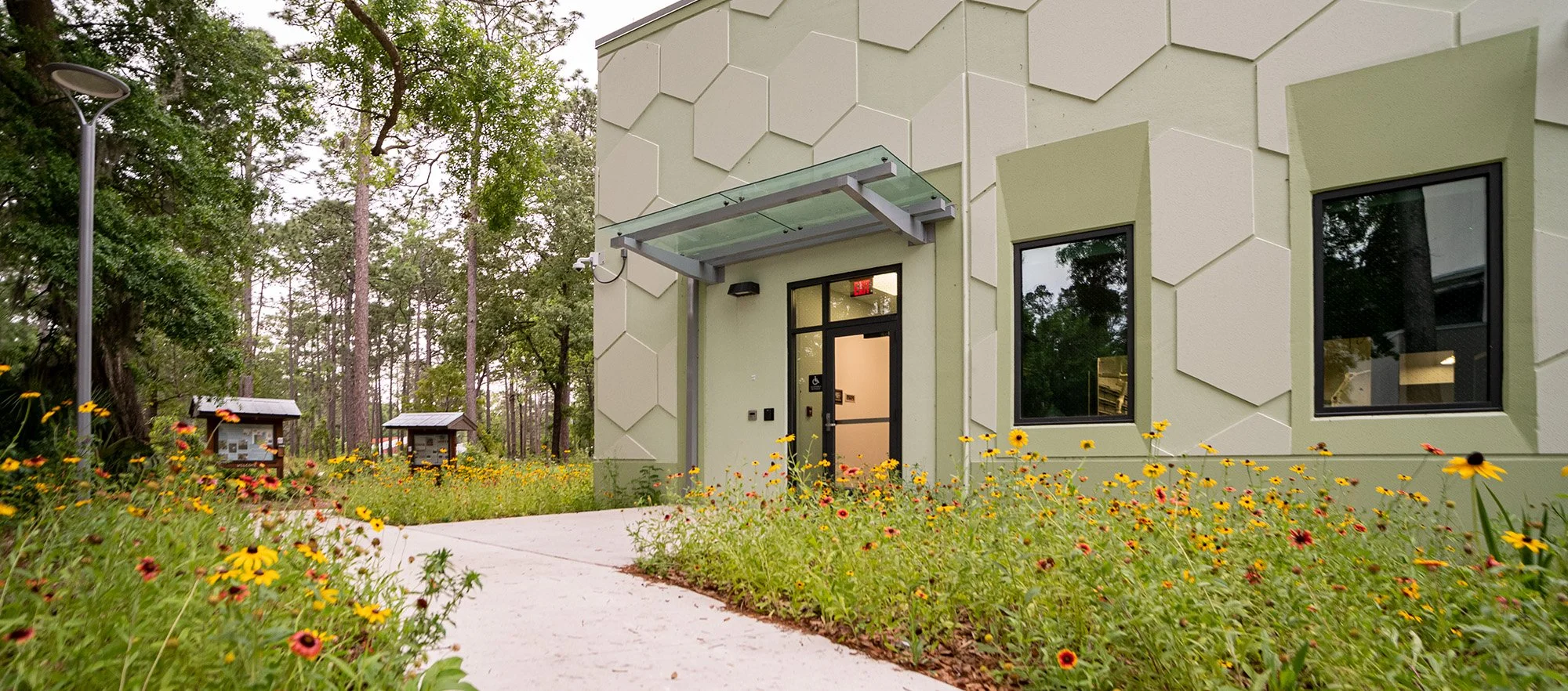 Exterior of a modern building with a hexagonal pattern on its facade, surrounded by a garden with yellow and red flowers, trees, and a narrow path leading to the entrance.