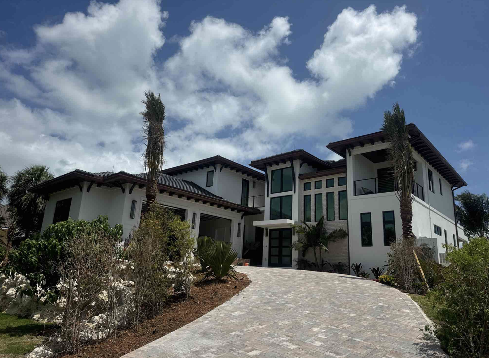 A modern two-story house with a white exterior, large windows, and a sloped dark roof, surrounded by palm trees and landscaped garden, under a partly cloudy sky.