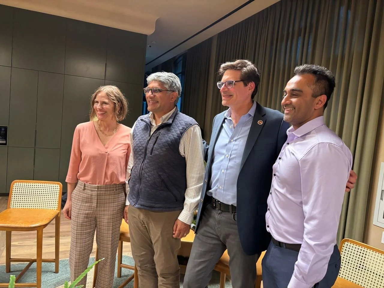 Four diverse people standing indoors, smiling, with chairs and curtains in the background.