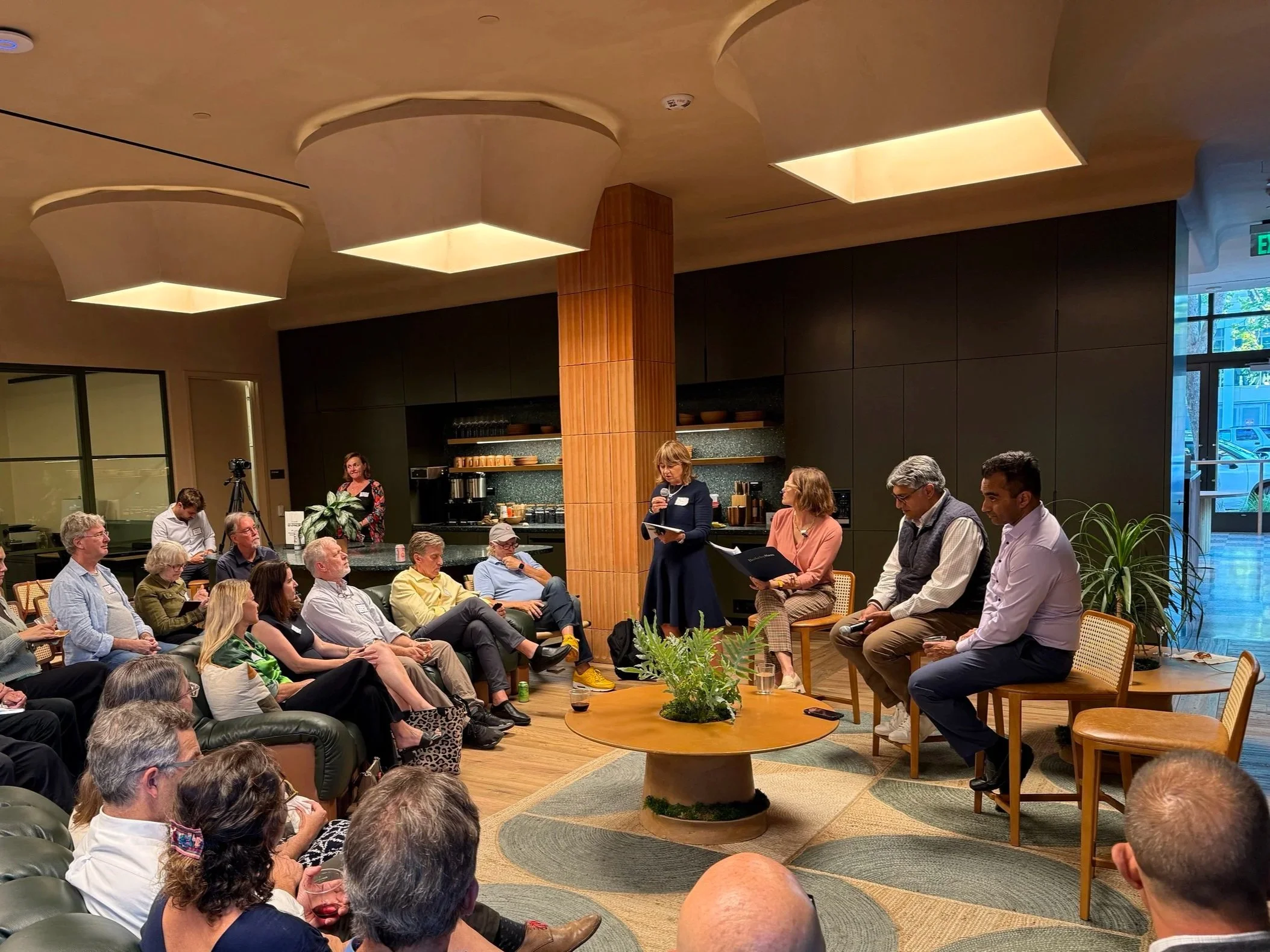 A group of people attending a panel discussion or presentation indoors, with some seated and some standing, featuring a woman speaking into a microphone and others seated beside her, in a modern room with warm lighting, plants, and a black and wooden interior design.