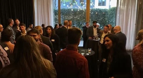 A group of people gathered at a networking event or reception indoors, standing around small tables, engaging in conversations, with large windows and curtains in the background.