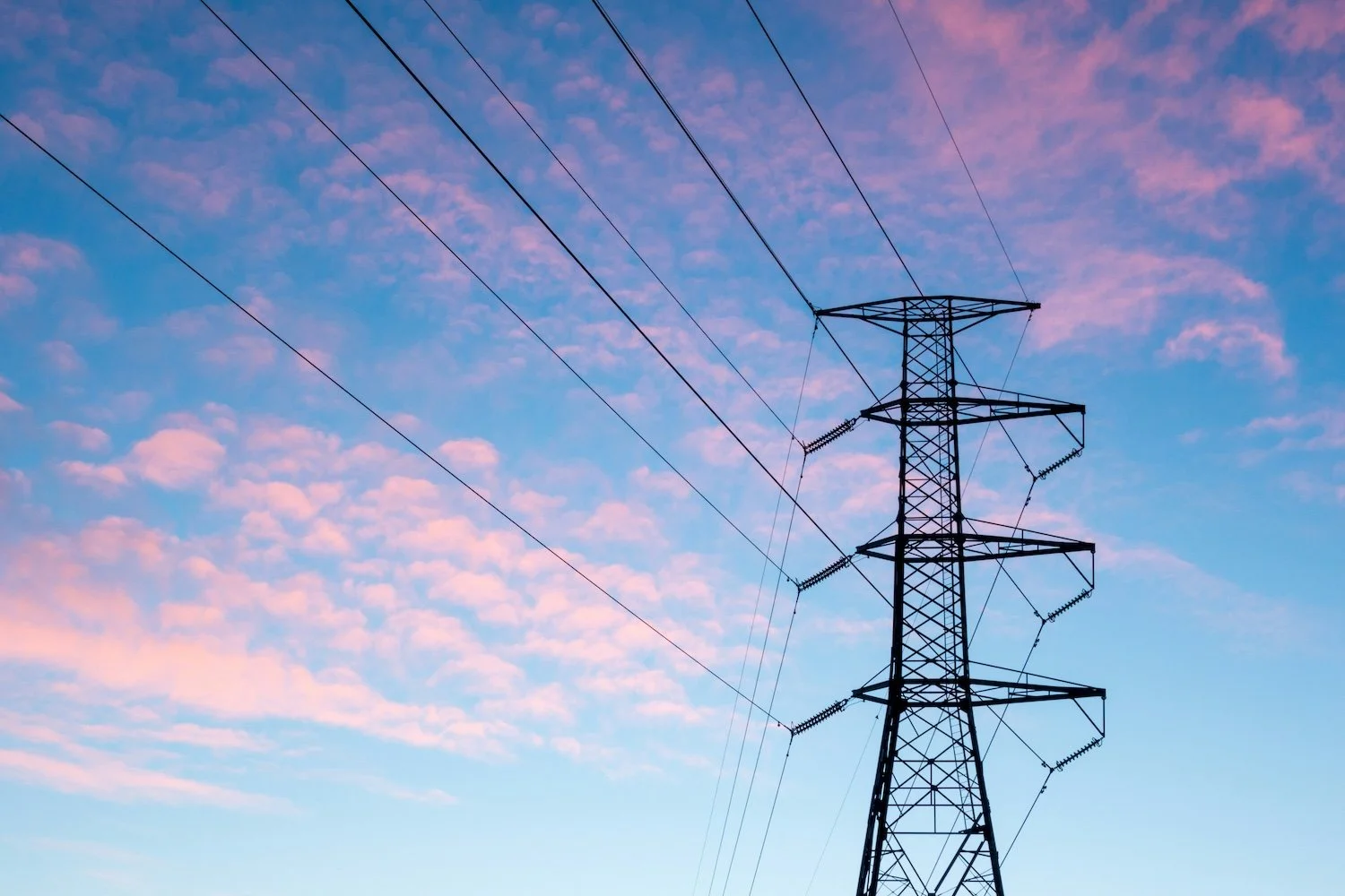A tall electrical transmission tower with power lines against a sky with pink clouds at sunset.