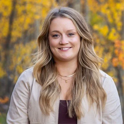 A woman with long, wavy blonde hair smiling outdoors with yellow autumn trees in the background.