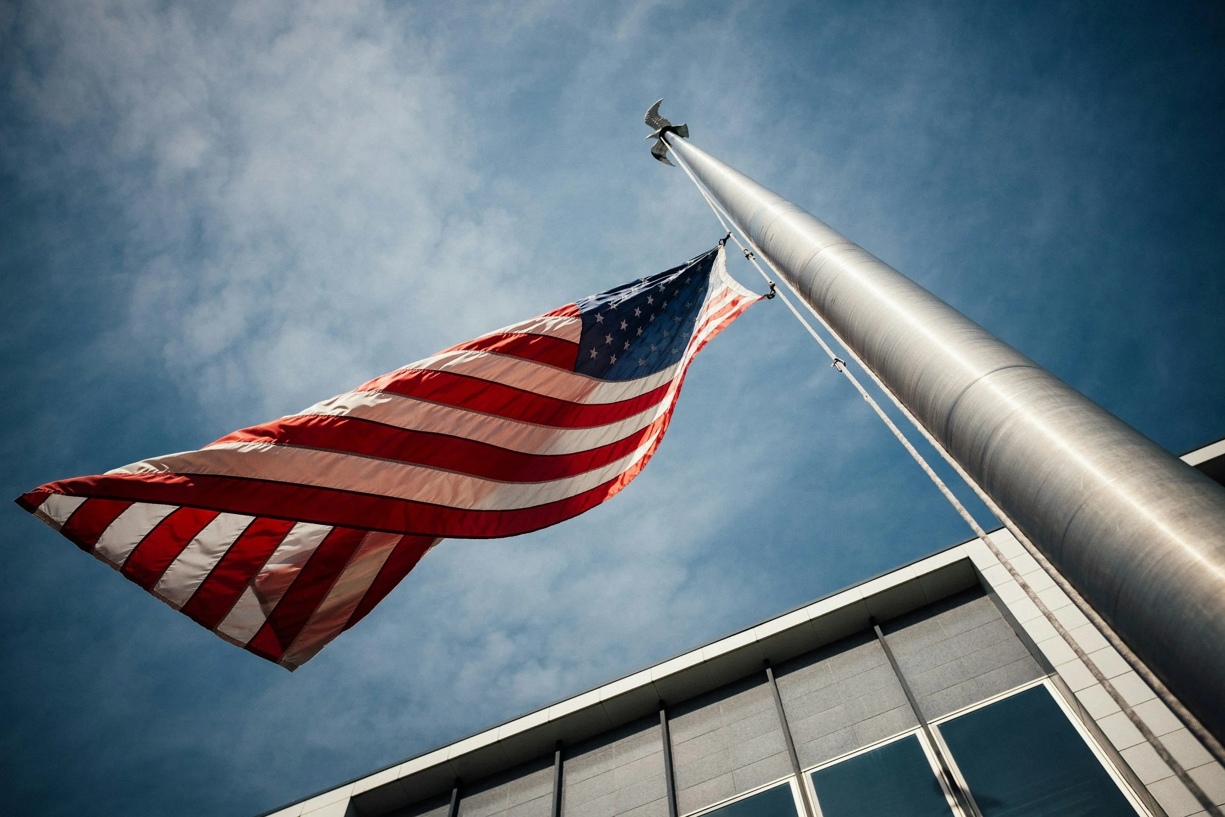 American flag flying at half-mast against a blue sky near a modern building.