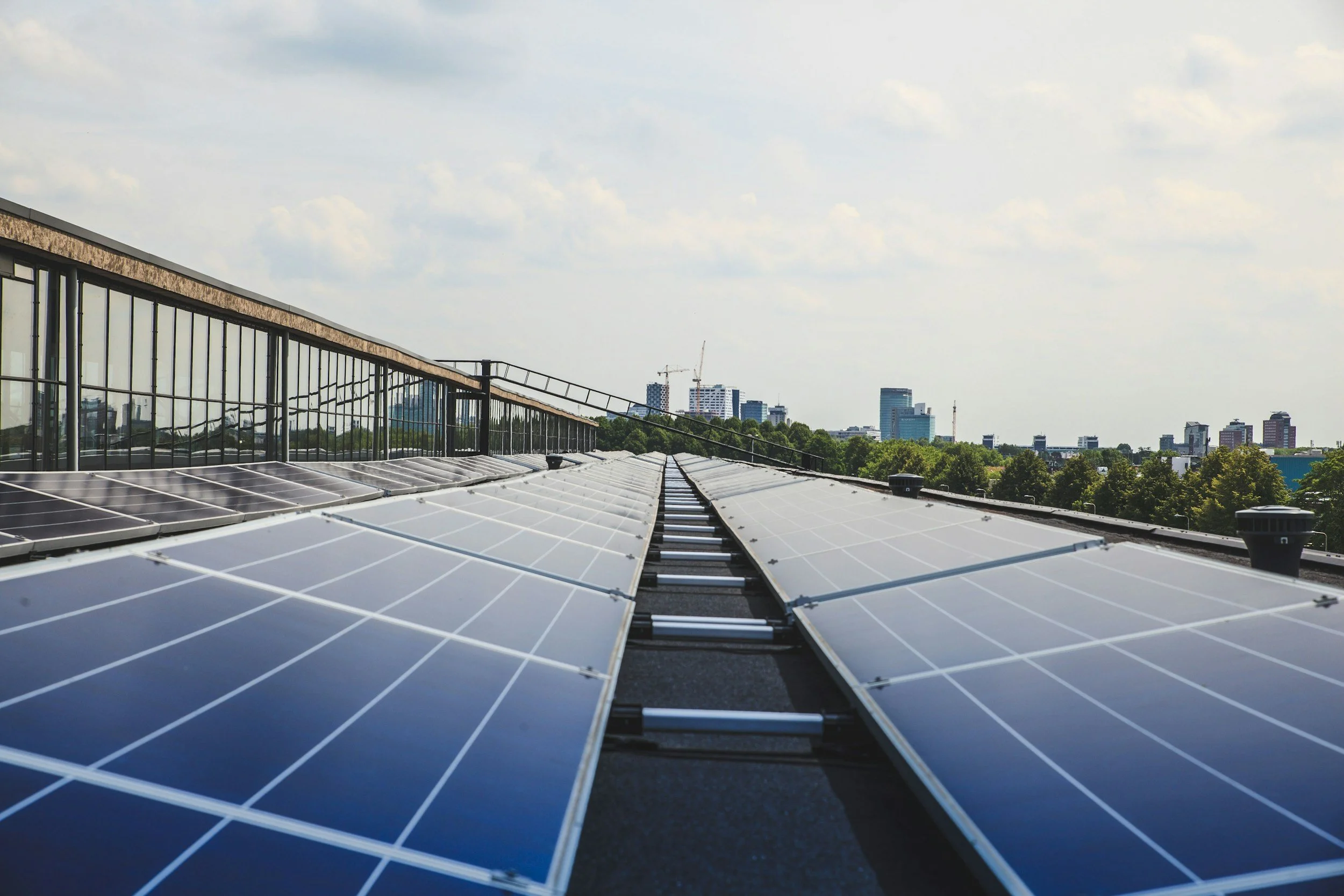 Rows of solar panels installed on a rooftop with a city skyline in the background under a partly cloudy sky.