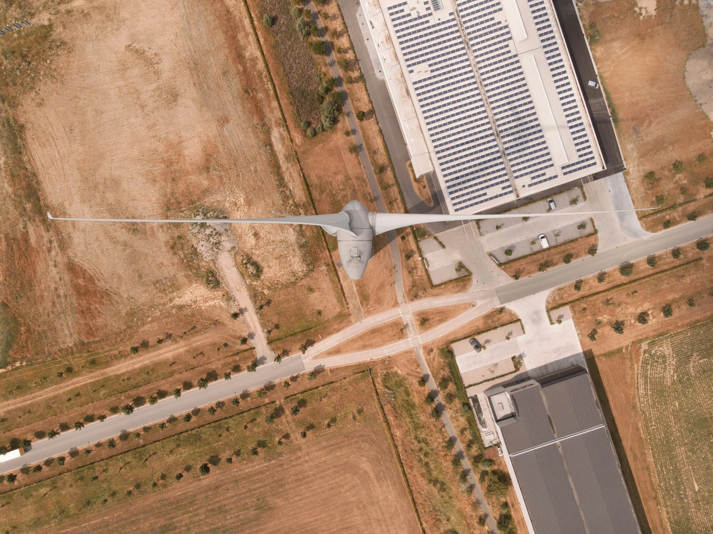 An aerial view of a wind turbine next to modern buildings with solar panels, surrounded by rural farmland and roads.
