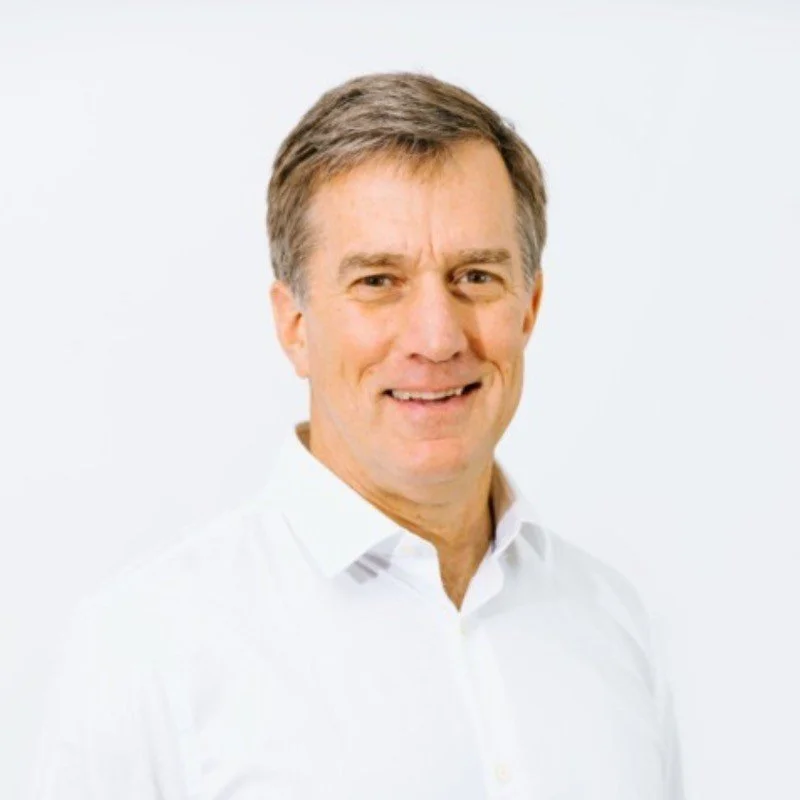 A middle-aged man with light skin, short brown hair, wearing a white button-up shirt, smiling against a plain white background.