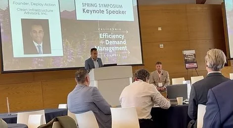 A speaker at a conference presentation standing at a podium with a PowerPoint slide behind him. The slide shows a photo of a man and text that reads 'Spring Symposium Keynote Speaker' and 'California Efficiency + Demand Management'. Several attendees are seated at tables facing the speaker.