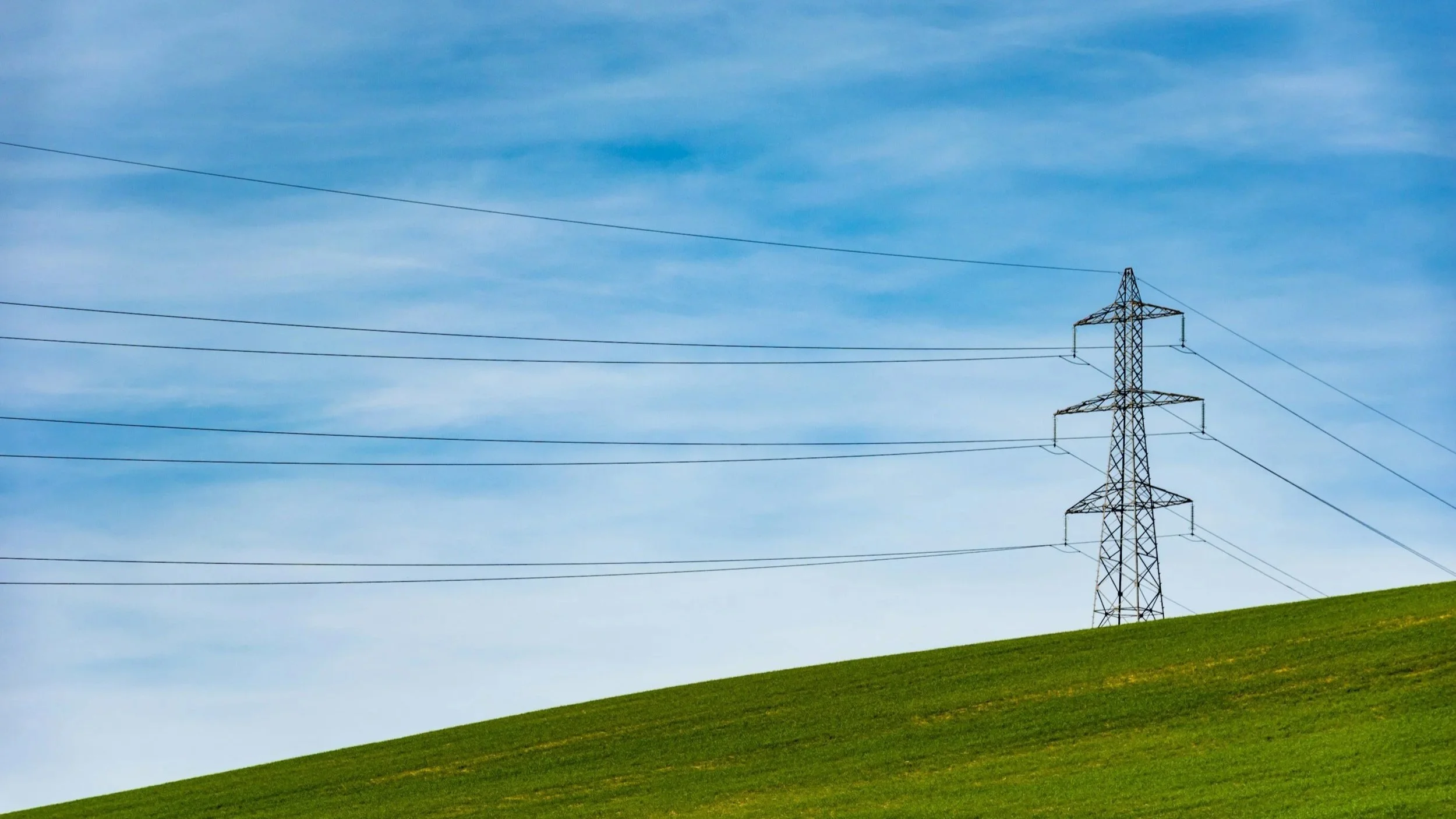 A green grassy hill with a tall electrical transmission tower and power lines against a blue sky with some clouds.