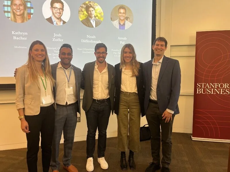 Five professionally dressed individuals standing together in front of a presentation screen and a Stanford Business banner in a conference room.