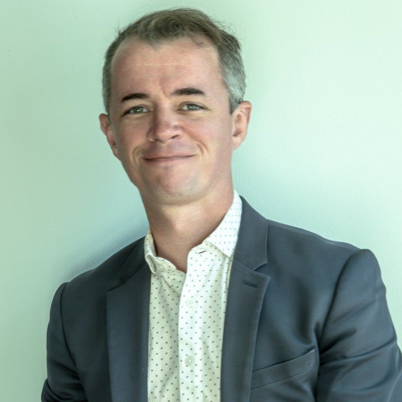 A smiling man in a dark blazer and a white dotted shirt, standing against a plain light-colored wall.