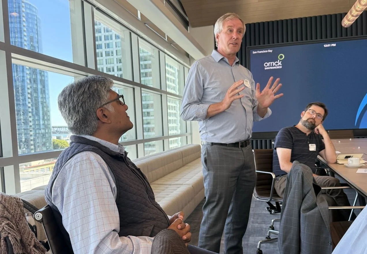 A man standing and speaking at a business meeting in a modern conference room with tall windows and city views. Two other men are seated, listening; one is looking up at the speaker, the other is resting his head on his hand.