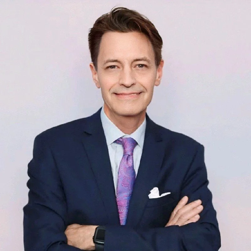Portrait of a man in a dark suit and purple tie, smiling with arms crossed, against a plain light background.