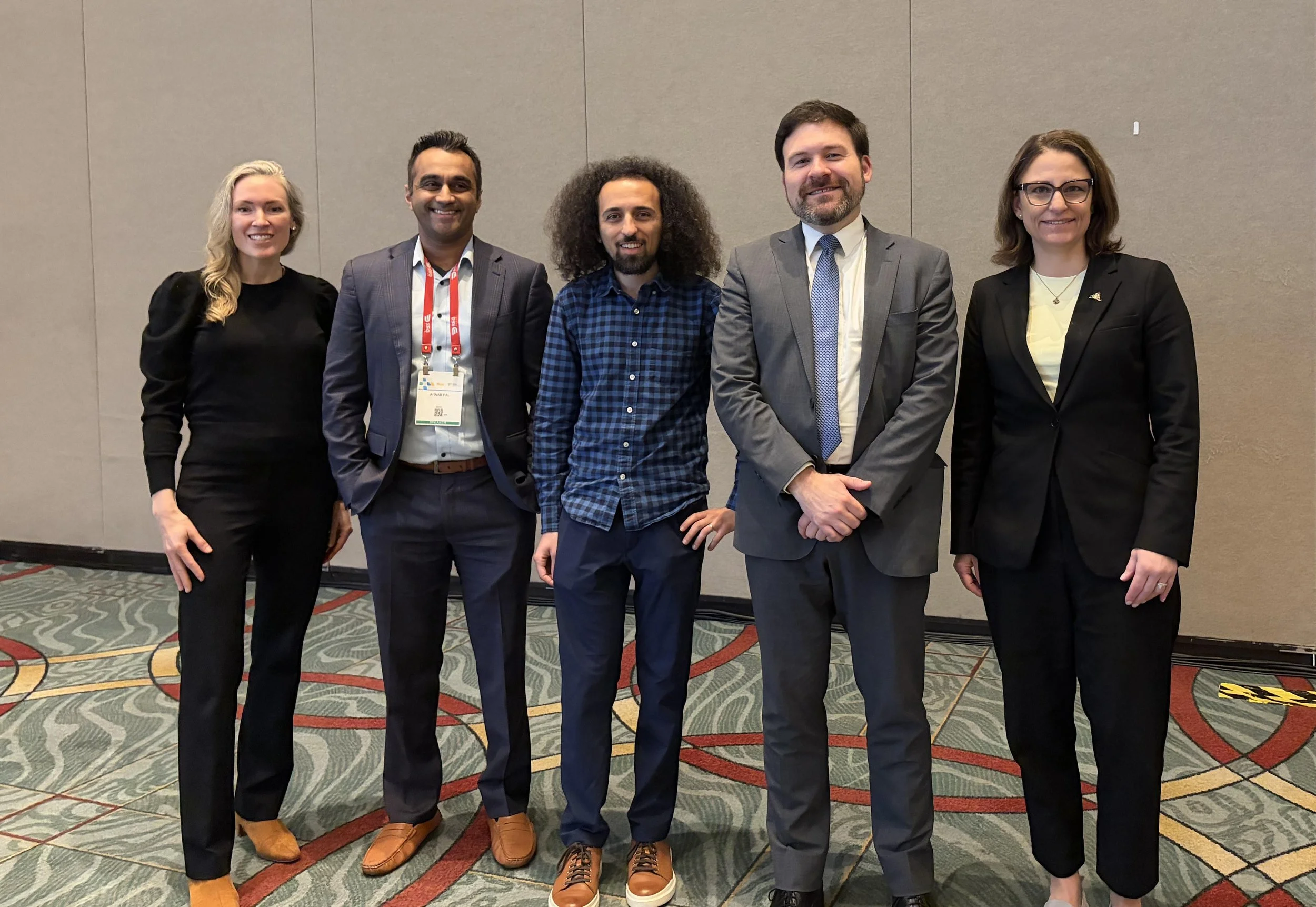 Group of five professionally dressed individuals, three men and two women, posing together indoors at a conference or event.