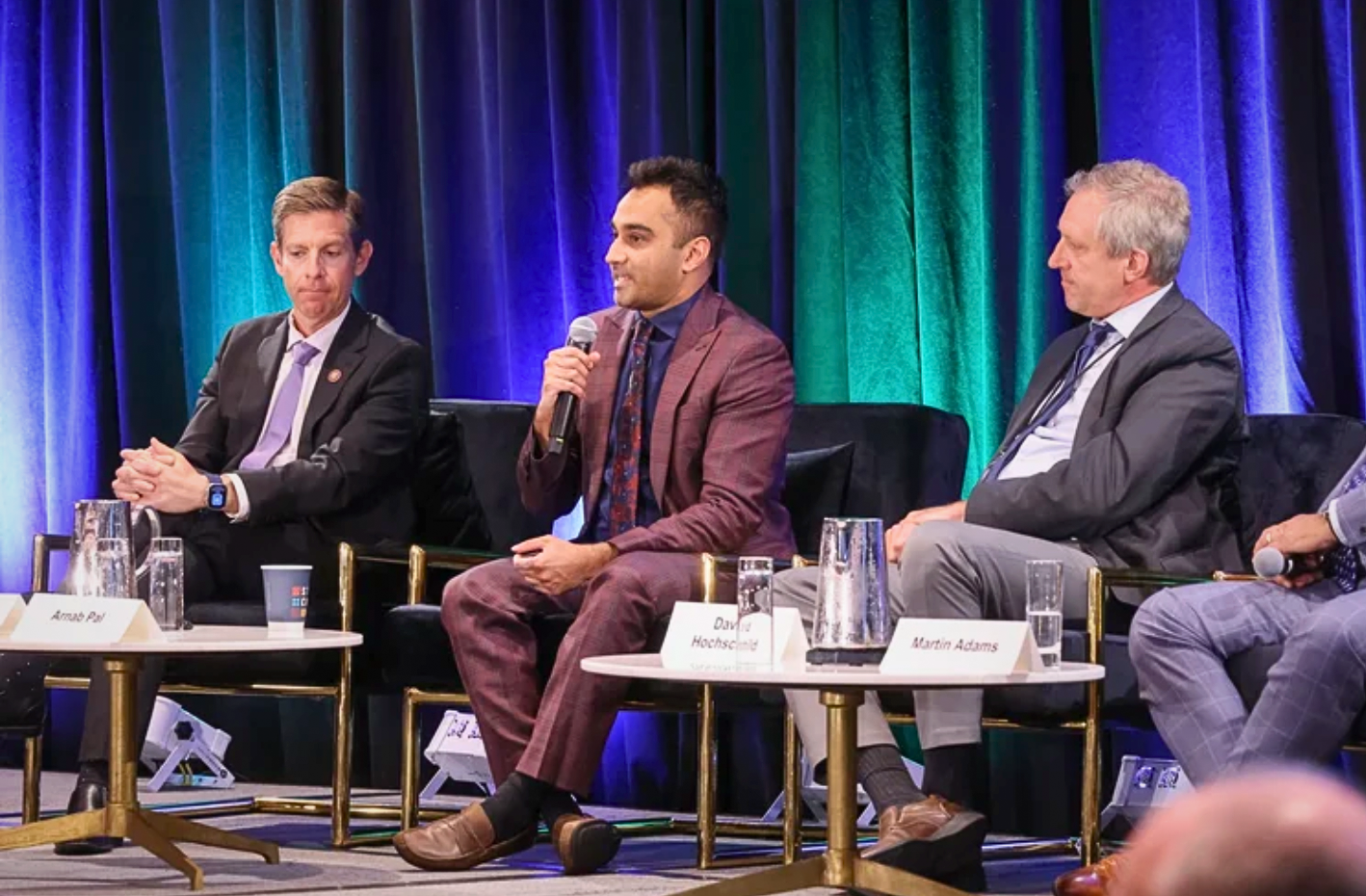 A panel discussion with three men seated on stage, one speaking into a microphone, in front of colorful curtains.
