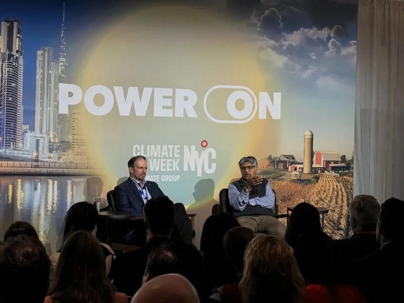 Two men speaking at a panel discussion during Climate Week NYC, with a backdrop that says 'POWER ON' and features cityscape and farm images.