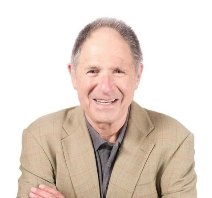 Portrait of a smiling elderly man with gray hair, wearing a light-colored blazer over a dark shirt, against a plain white background.