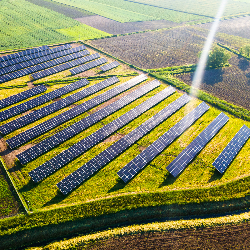 Aerial view of solar panels installed on a farmland field with patches of green and brown earth, and a sunbeam shining down.