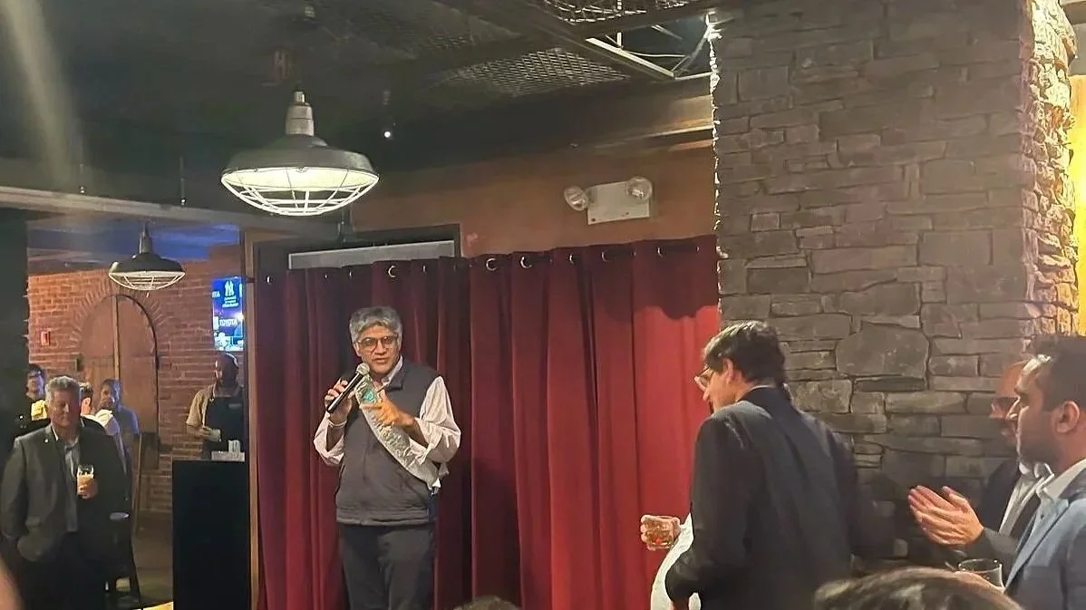 A woman with short gray hair, glasses, and a gray vest over a white shirt speaking into a microphone at a social gathering inside a bar or restaurant. Several people are standing nearby, some clapping and holding drinks. The setting has brick walls, red curtains, and industrial-style lighting.