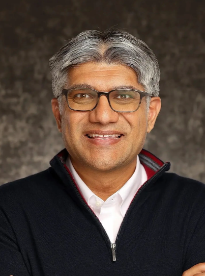 A middle-aged man with gray hair and glasses smiling, wearing a black zip-up sweater over a white collared shirt, standing against a dark textured background.