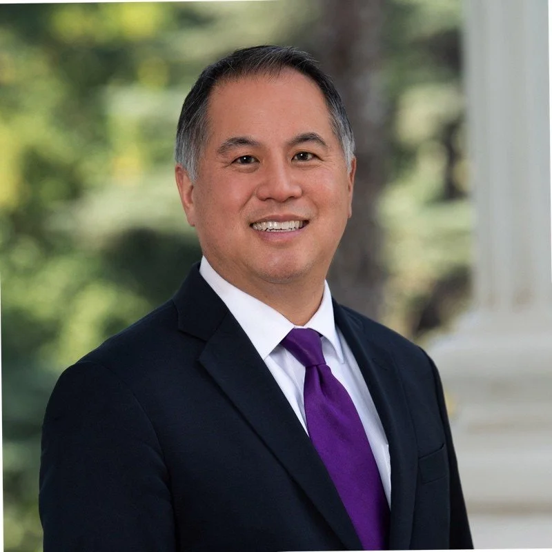 A professional man dressed in a black suit, white shirt, and purple tie, smiling, with a blurred outdoor background.