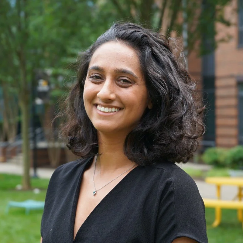 A woman with curly dark hair smiling outside in a park with trees in the background and a yellow bench.