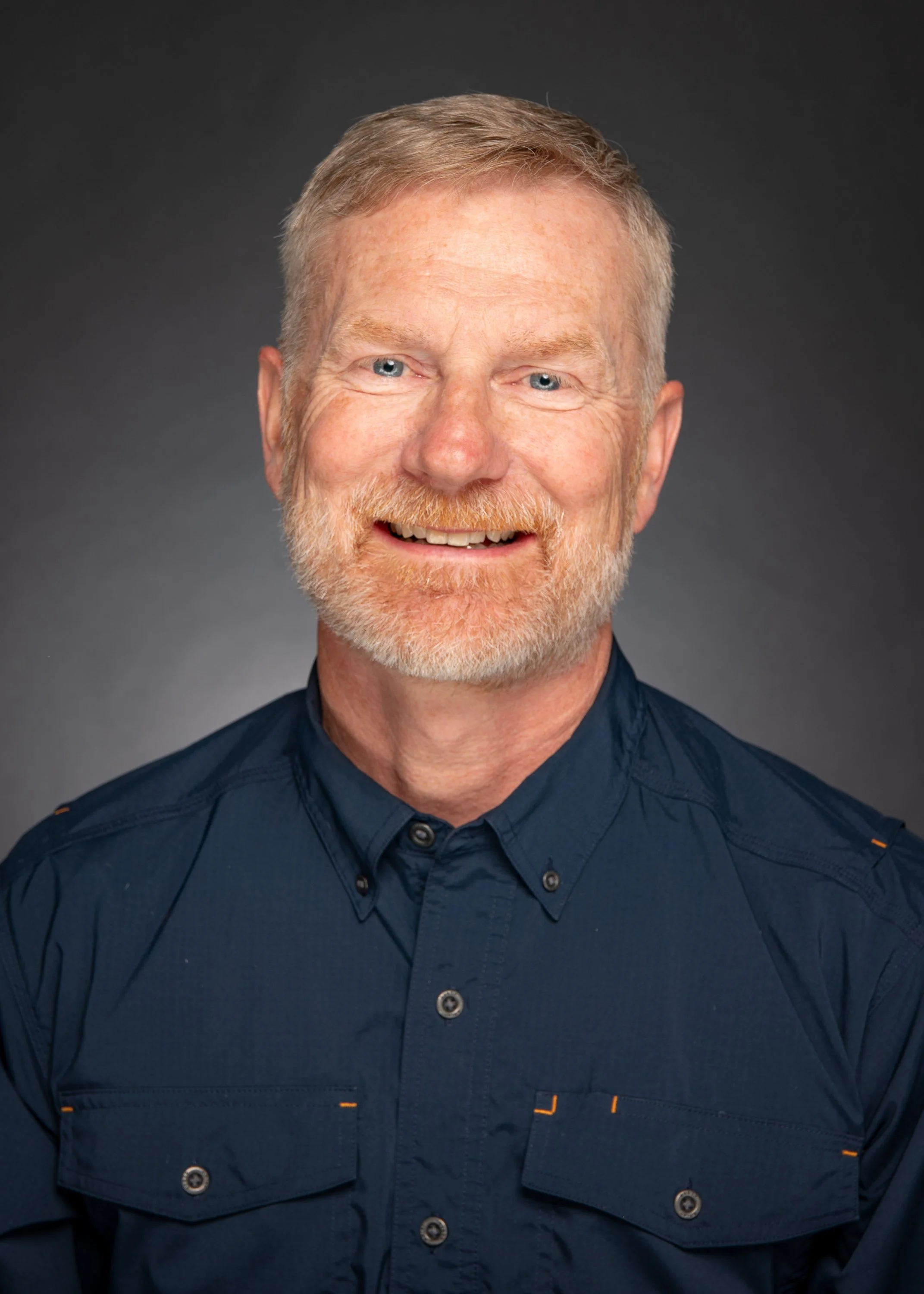 Close-up portrait of a smiling middle-aged man with light hair, beard, and blue eyes, wearing a dark blue button-up shirt, against a dark gray background.