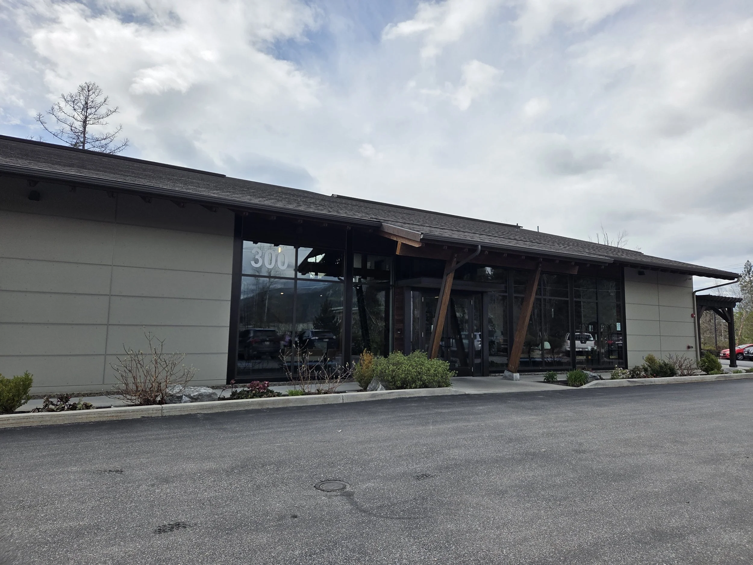 Exterior view of a modern commercial building with large glass windows, a sloped roof, and landscaping in the front. The sky is cloudy.