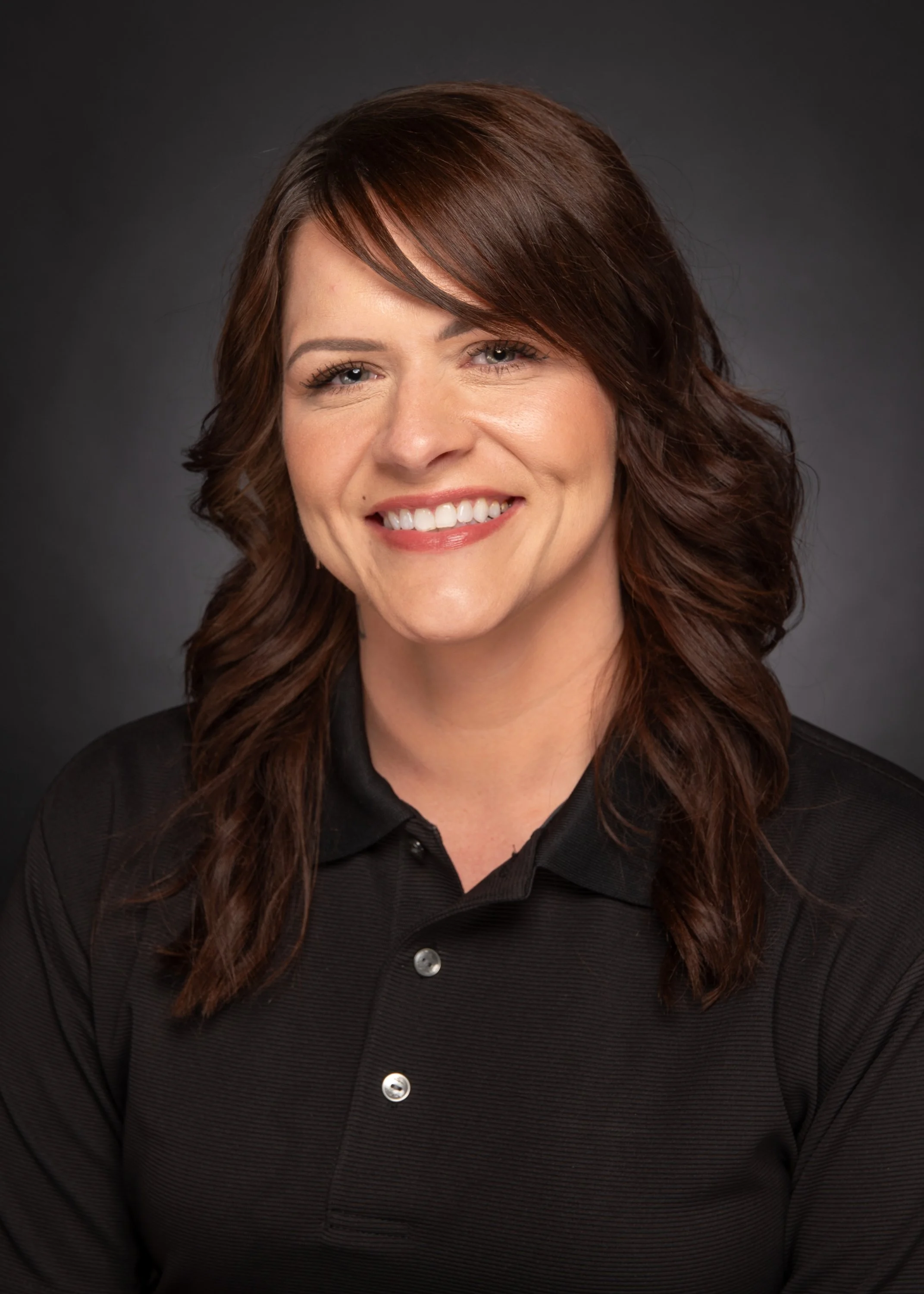 Portrait of a woman with shoulder-length wavy brown hair, smiling, wearing a black polo shirt, against a dark gray background.