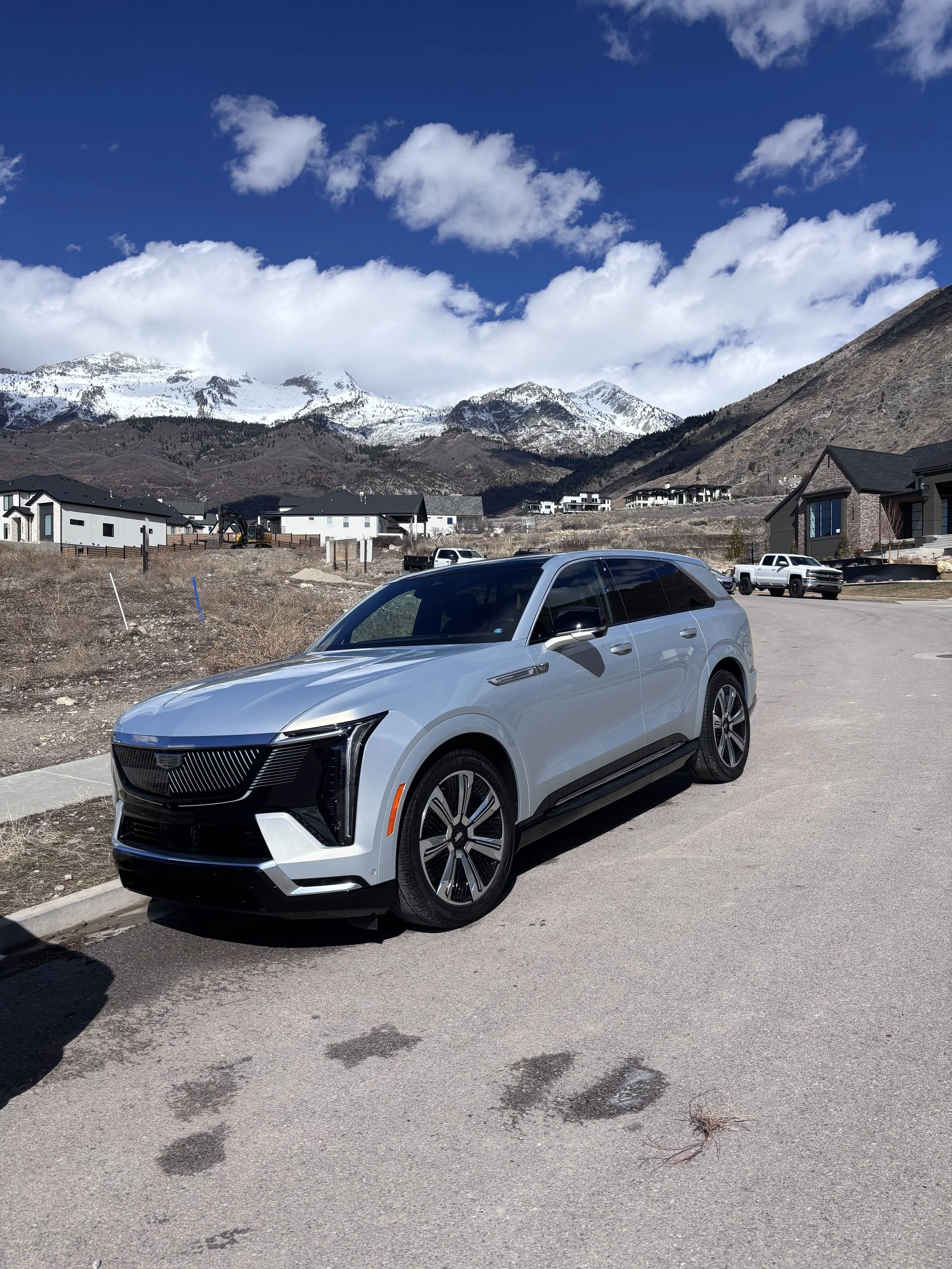 A modern silver SUV parked on a paved street with snow-capped mountains and partly cloudy blue sky in the background.