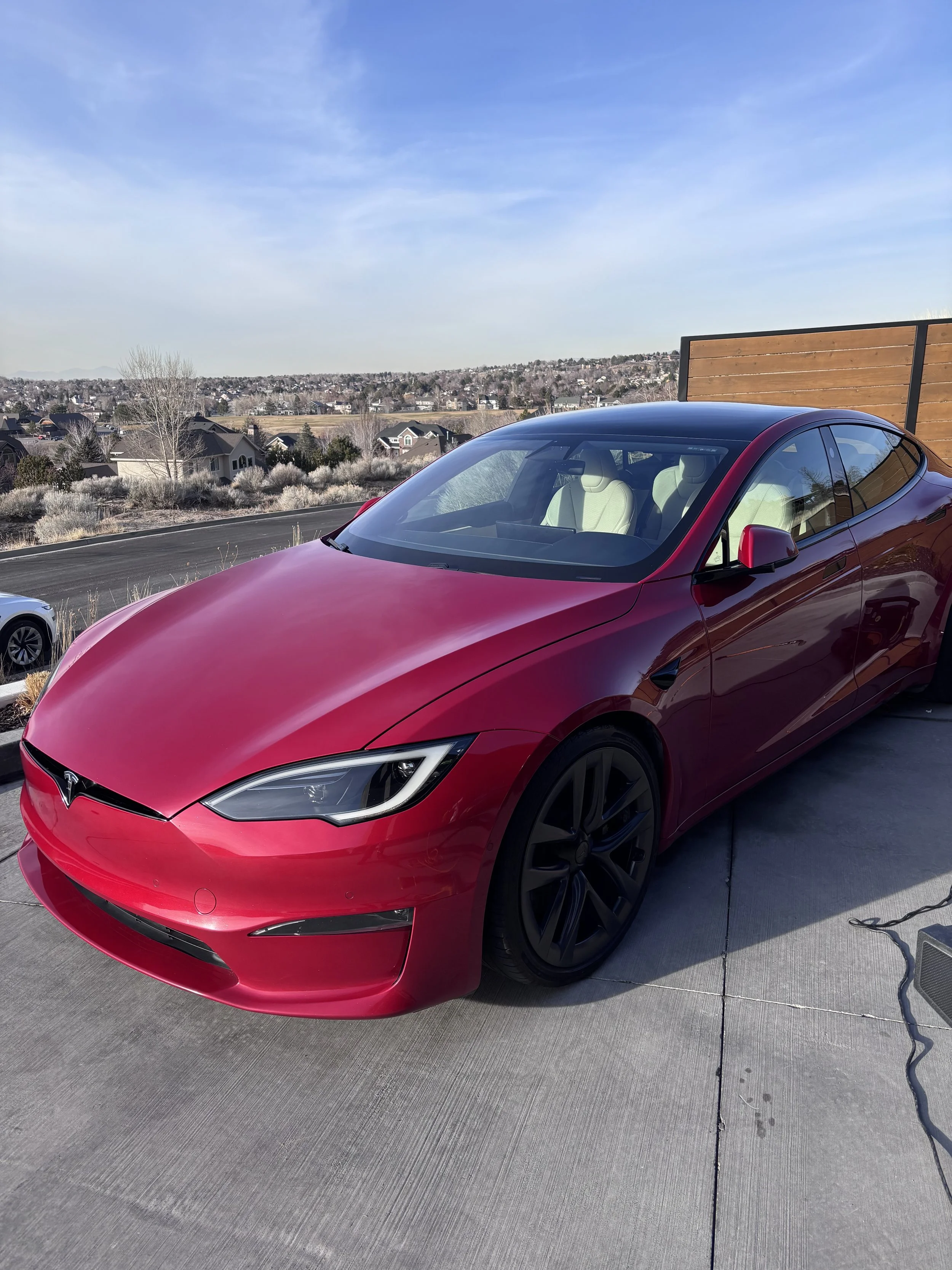 Red Tesla Model S parked on a driveway with a scenic suburban landscape in the background, clear blue sky, and a wooden fence