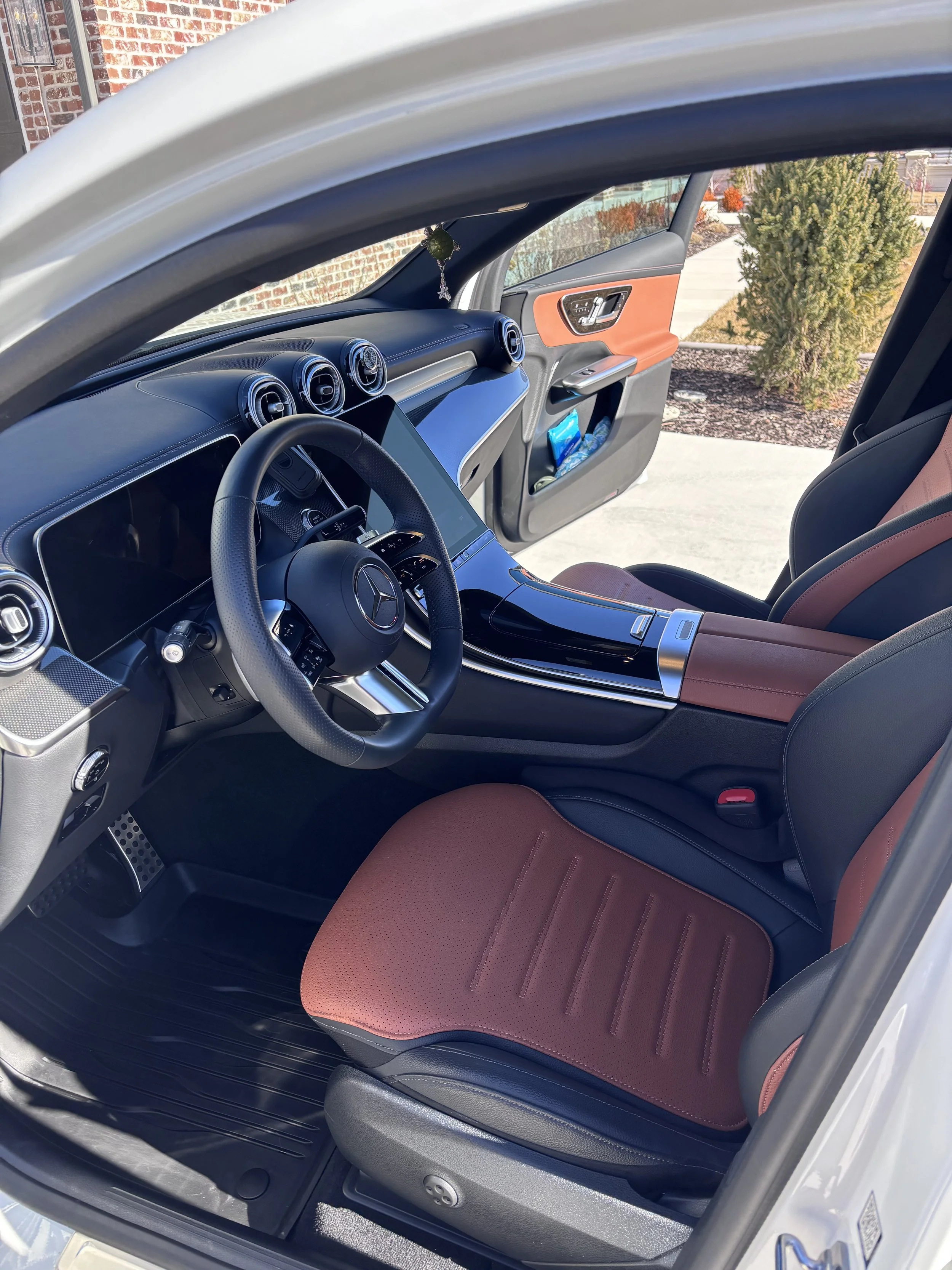 Interior of a Mercedes-Benz vehicle showing the steering wheel, dashboard with three circular air vents, a digital display, and tan and black seats with red accents. Part of the open door and outdoor background with a shrub and sidewalk are visible.