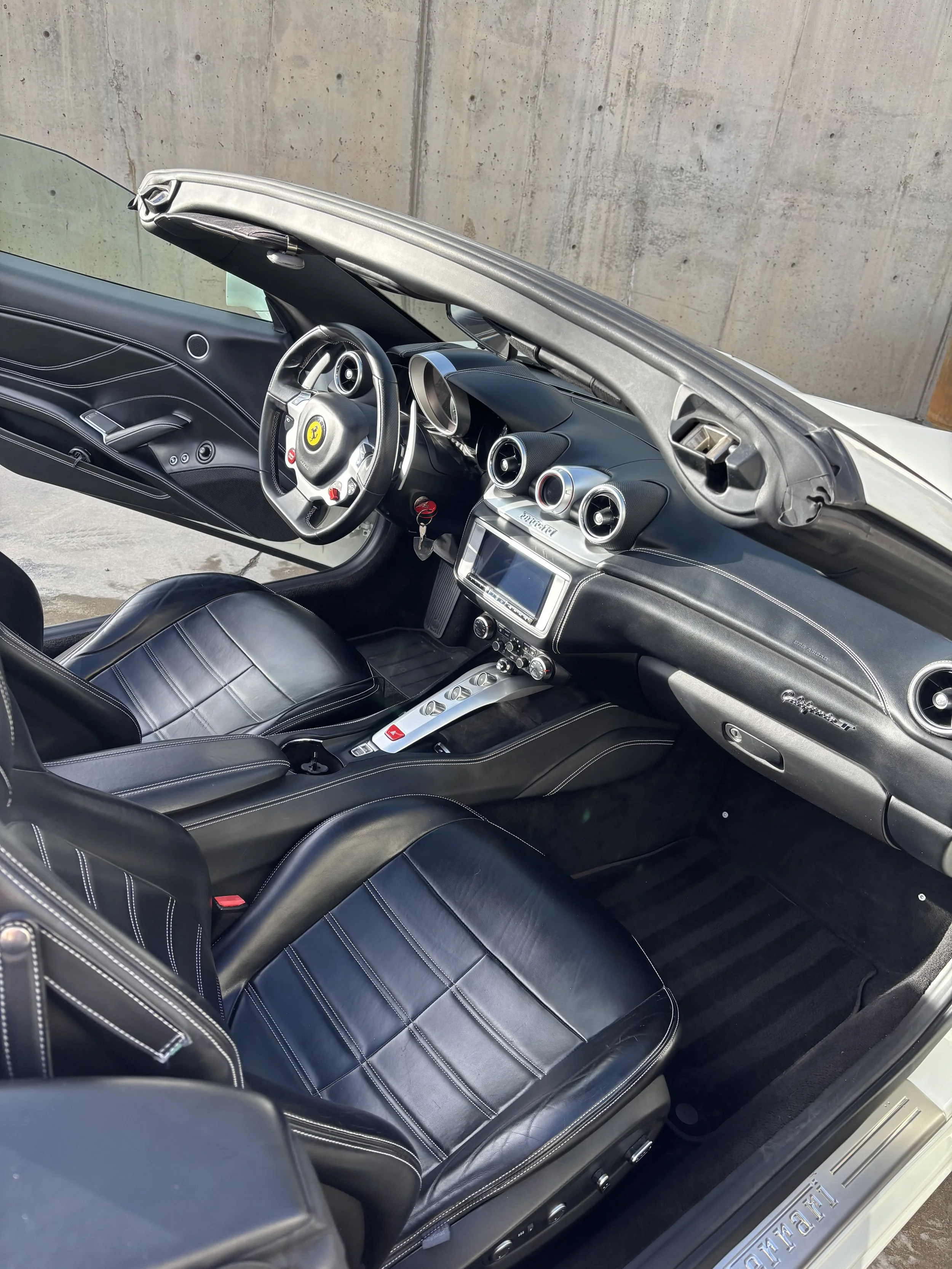 Interior view of a black Ferrari convertible with leather seats and a modern dashboard featuring circular air vents and a touchscreen display.