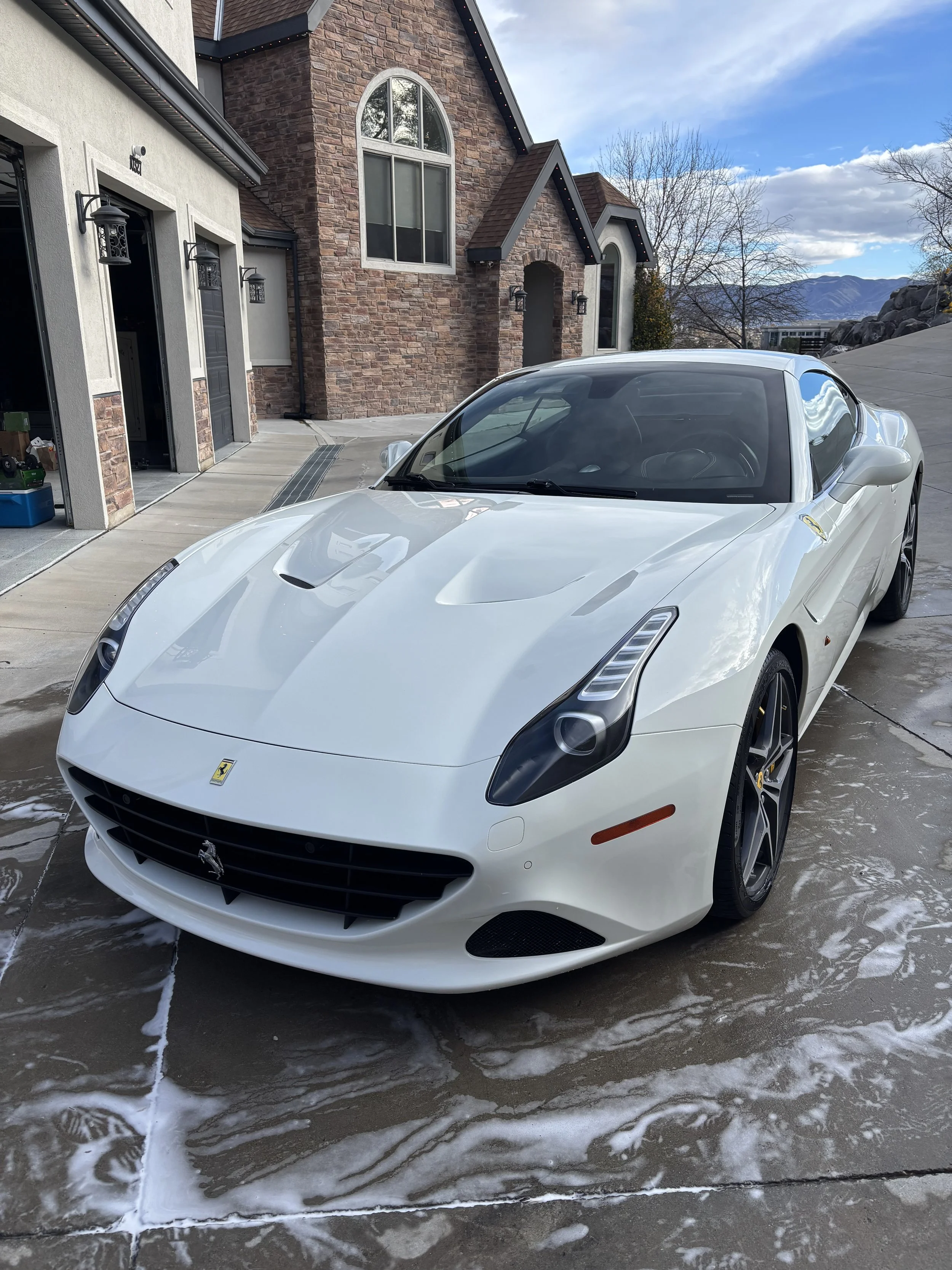 White Ferrari sports car parked on driveway outside a house with a brick and stucco exterior, arched windows, and mountain view in the background.